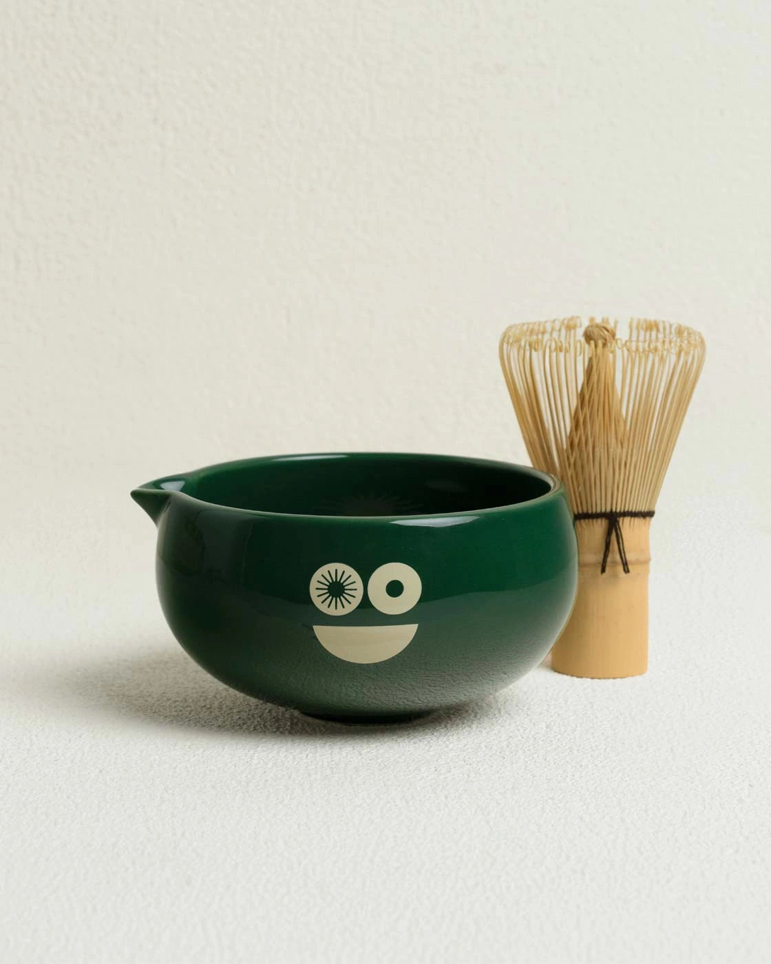 Green ceramic bowl with a face design and bamboo whisk on a beige background
